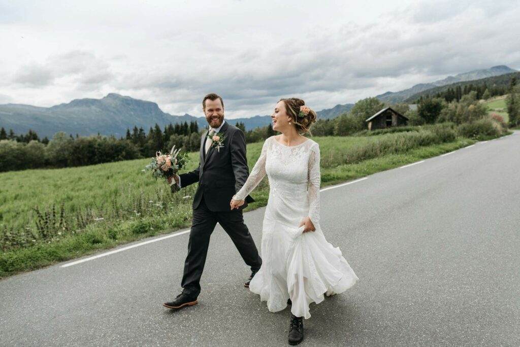 Bride and groom holding hands and walking on the road, with a green field and the Dolomite mountains in the background.