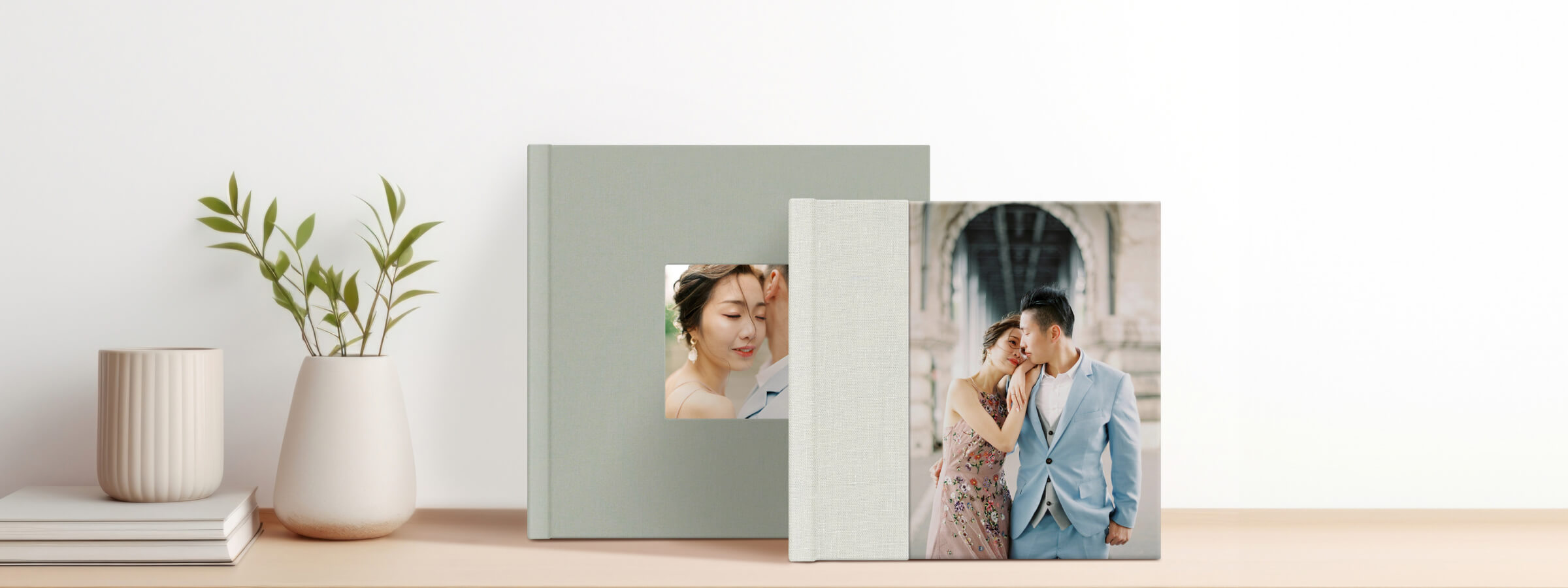 two photo albums in pastel colors featuring photos of a couple dressed up, stand on a table next to a minimalistic vase with greens, a coffee mug and some magazines