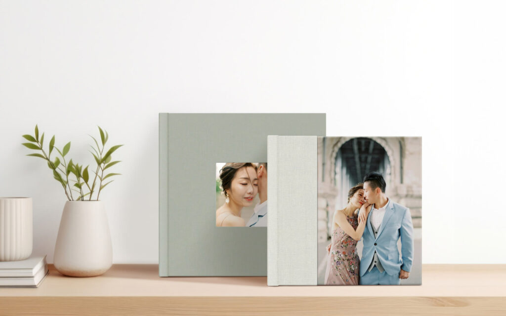 two pastel photo albums stand on a table, next to a vase with a leafy plant. The albums are in pastel colors and feature photos of a bride and groom hugging